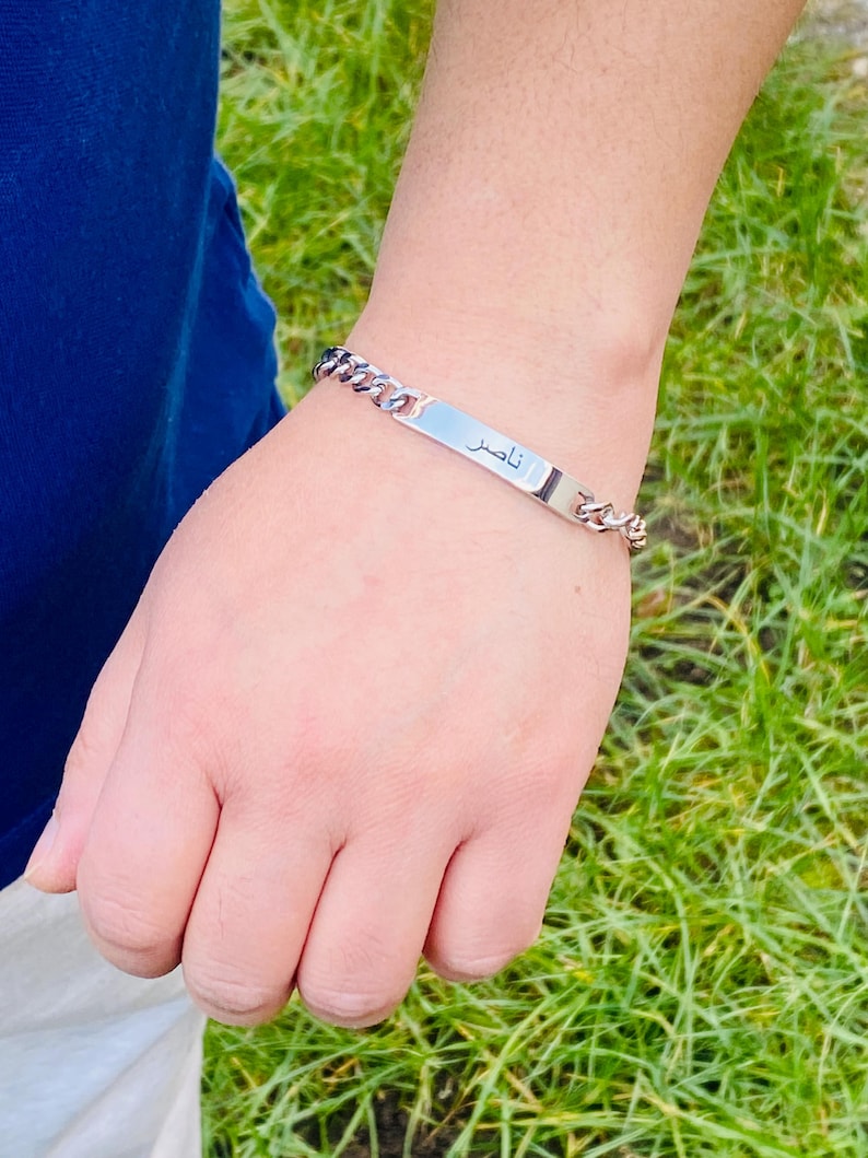 May include: A silver-toned metal chain link bracelet featuring a flat, rectangular identification plate in the center. The plate is engraved with the Arabic name "ناصر" in black lettering. The bracelet has a polished finish and a secure clasp.