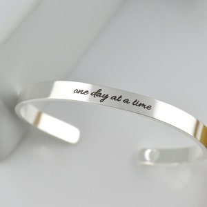 May include: A polished silver-tone cuff bracelet featuring the engraved black text "one day at a time" in a cursive script font. The bracelet has an open-ended design with rounded terminals and a smooth, reflective metallic surface finish.