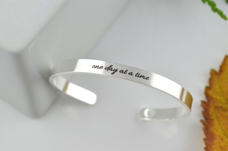 May include: A polished silver-tone cuff bracelet featuring the engraved black text "one day at a time" in a cursive script font. The bracelet has an open-ended design with rounded terminals and a smooth, reflective metallic surface finish.
