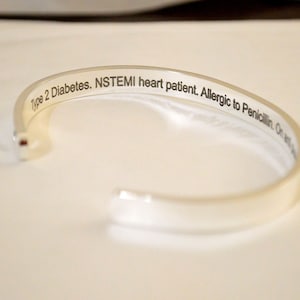 May include: A silver-colored metal cuff bracelet with an open design. The interior surface features black engraved text that reads: Type 2 Diabetes. NSTEMI heart patient. Allergic to Penicillin. The bracelet has a smooth, polished exterior finish.