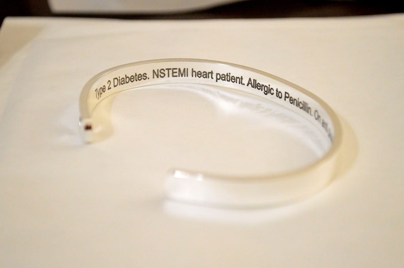 May include: A silver-colored metal cuff bracelet with an open design. The interior surface features black engraved text that reads: Type 2 Diabetes. NSTEMI heart patient. Allergic to Penicillin. The bracelet has a smooth, polished exterior finish.