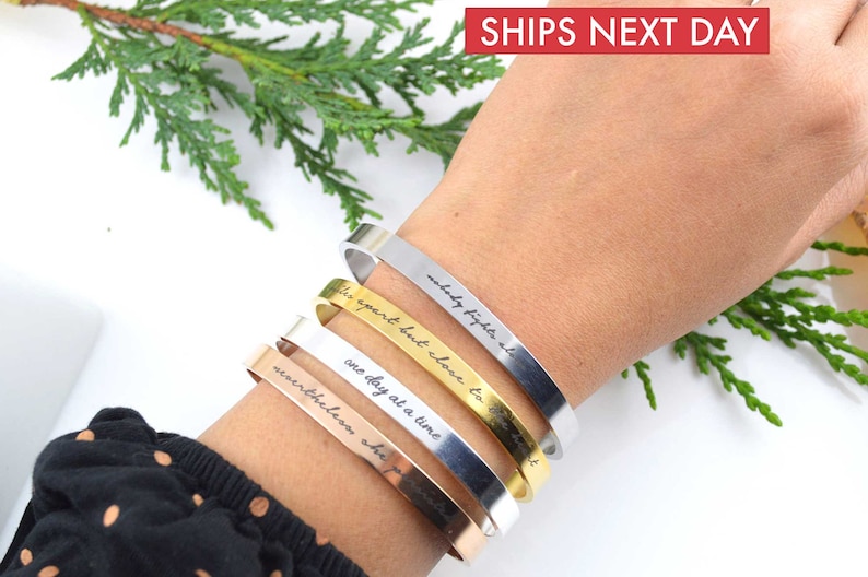 May include: A set of four metal cuff bracelets in silver, gold, and rose gold tones. Each bracelet features a smooth, flat surface with engraved black cursive text. The inscriptions include phrases such as nobody fights alone, one day at a time, and nevertheless she persisted.