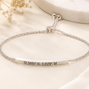Op de afbeelding: Een verstelbare zilverkleurige armband met een dunne, gebogen metalen staaf gegraveerd met de coördinaten 51.5014° N, 0.1419° W. De armband is gemaakt van een fijne ketting met een schuifkraalmechanisme voor het aanpassen van de maat.