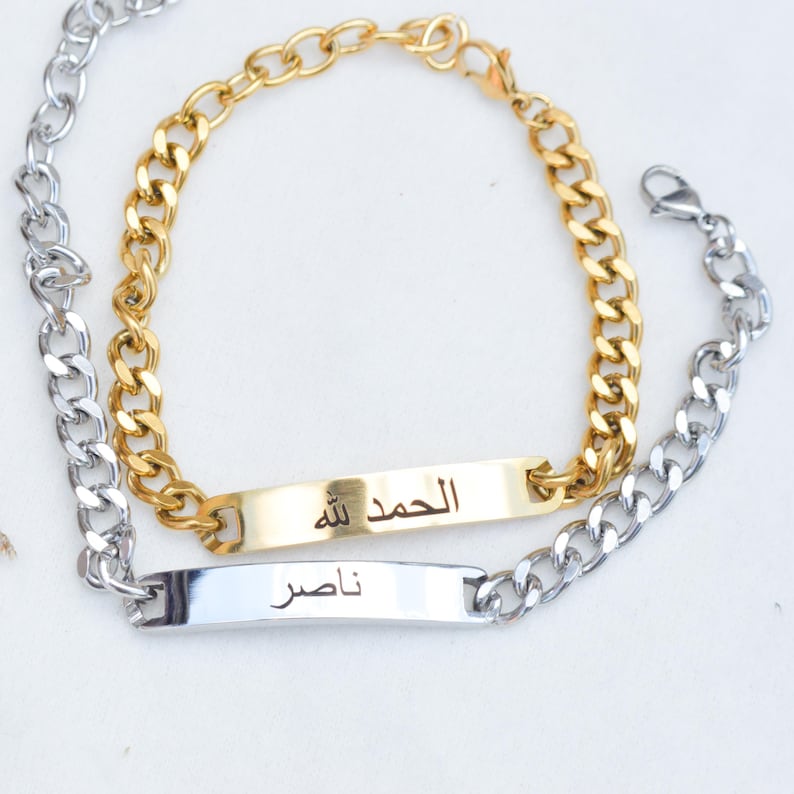 May include: Two curb chain bracelets with rectangular metal ID plates. One bracelet is gold-toned with the Arabic text "الحمد لله" engraved on the plate. The other bracelet is silver-toned with the Arabic text "ناصر" engraved on the plate.
