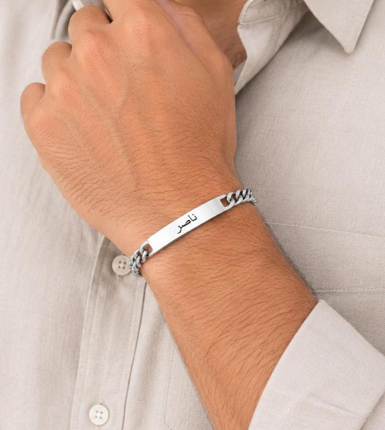 May include: A silver-toned metal chain bracelet featuring a flat, rectangular identification plate in the center. The plate is engraved with the Arabic name Nasser in black lettering. The bracelet consists of interlocking curb chain links.