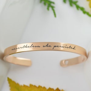 May include: A rose gold-toned metal cuff bracelet with a smooth, polished finish. The outer surface features the engraved black text "nevertheless, she persisted." in a cursive script font. The bracelet has an open-ended design with rounded ends.