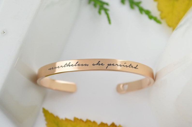 May include: A rose gold-toned metal cuff bracelet with a smooth, polished finish. The outer surface features the engraved black text "nevertheless, she persisted." in a cursive script font. The bracelet has an open-ended design with rounded ends.