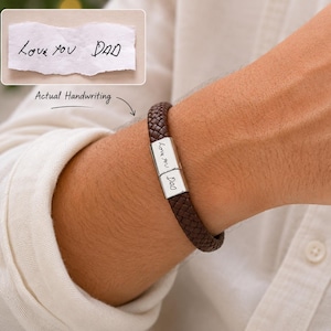 May include: A dark brown braided leather bracelet featuring a rectangular silver-toned metal clasp. The clasp is engraved with the handwritten text Love you DAD, which matches the handwriting shown on a small piece of paper in the corner.