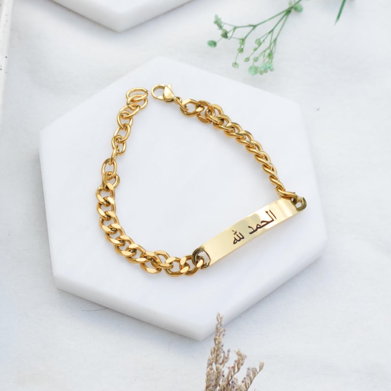 May include: A gold-toned chain link bracelet featuring a central rectangular metal plate engraved with the Arabic script text Alhamdullilah. The bracelet includes a lobster claw clasp closure and a series of interlocking oval links in a polished finish.