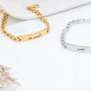 May include: Two chain link bracelets with rectangular identification plates. One bracelet is gold-toned with the Arabic text "الحمد لله" engraved on the plate. The other bracelet is silver-toned with the Arabic text "ناصر" engraved on the plate.