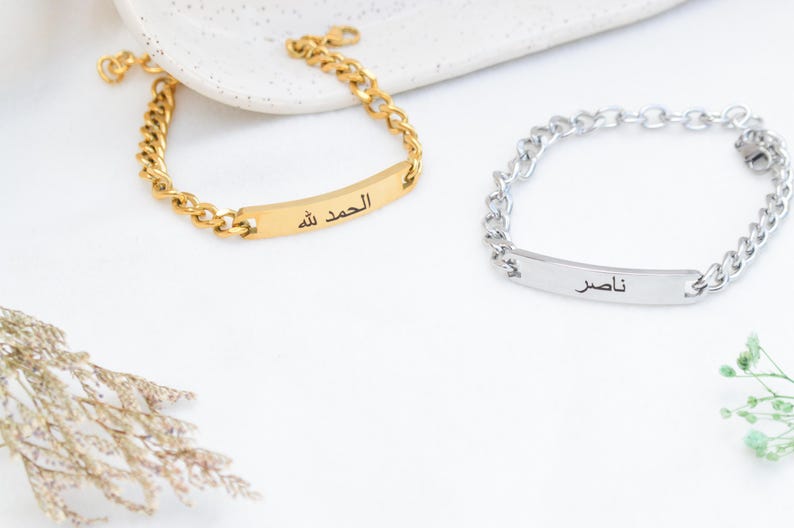 May include: Two chain link bracelets with rectangular identification plates. One bracelet is gold-toned with the Arabic text "الحمد لله" engraved on the plate. The other bracelet is silver-toned with the Arabic text "ناصر" engraved on the plate.