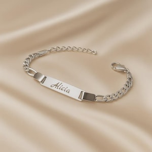 May include: A silver-toned metal chain bracelet featuring a central rectangular identification plate engraved with the name Alicia in a cursive script font. The bracelet includes a lobster claw clasp and a series of interlocking oval links.