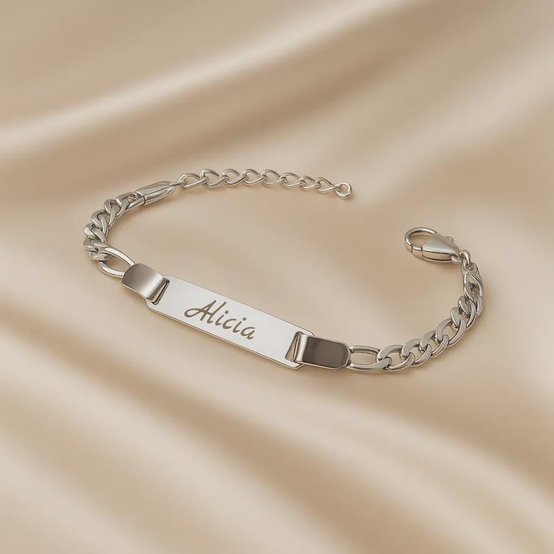 May include: A silver-toned metal chain bracelet featuring a central rectangular identification plate engraved with the name Alicia in a cursive script font. The bracelet includes a lobster claw clasp and a series of interlocking oval links.