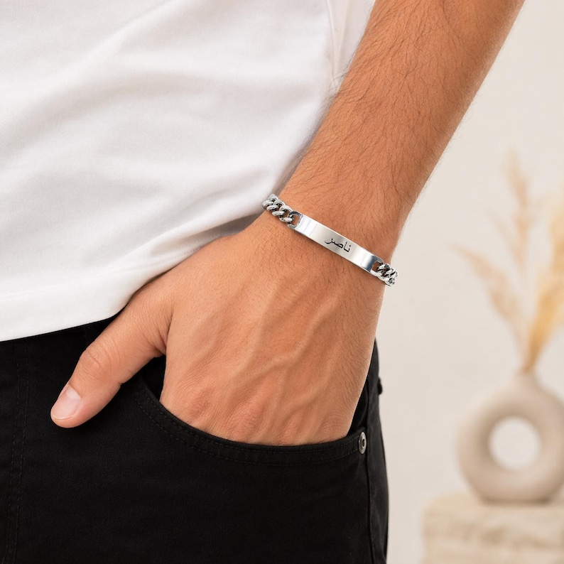 May include: A silver-toned metal chain bracelet featuring a flat, rectangular identification plate in the center. The plate is engraved with the Arabic name "ناصر" in black lettering. The bracelet consists of interlocking curb chain links.