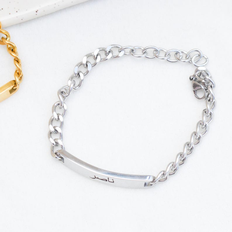 May include: A silver-toned chain link bracelet featuring a smooth, rectangular identification plate. The plate is engraved with the Arabic name "ناصر" in black lettering. The bracelet includes a lobster claw clasp and a short extender chain.