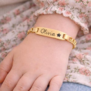 May include: A gold-toned metal identification bracelet featuring a flat rectangular center plate engraved with the name Olivia and a small star symbol, connected to a polished curb chain link band with a secure clasp mechanism.