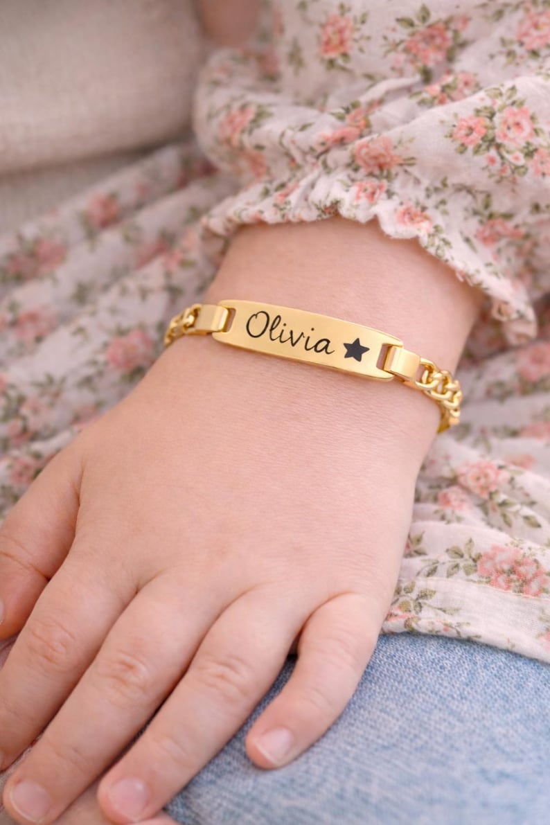 May include: A gold-toned metal identification bracelet featuring a flat rectangular center plate engraved with the name Olivia and a small star symbol, connected to a polished curb chain link band with a secure clasp mechanism.