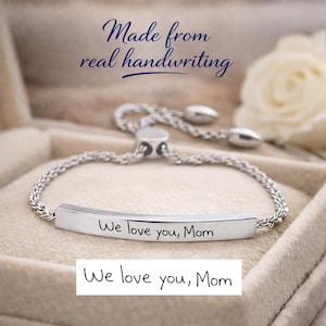 Peut inclure: Un bracelet réglable argenté doté d'une barre métallique incurvée gravée du texte "We love you, Mom" en écriture manuscrite. Le bracelet possède une chaîne torsadée et un fermoir coulissant. Le texte au-dessus indique "Made from real handwriting."