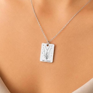 Può includere: Una collana d'argento con un ciondolo rettangolare. Il ciondolo presenta un'illustrazione in bianco e nero di due mani che si tendono l'una verso l'altra, con stelle sopra. Il testo "THE LOVERS" è sotto.