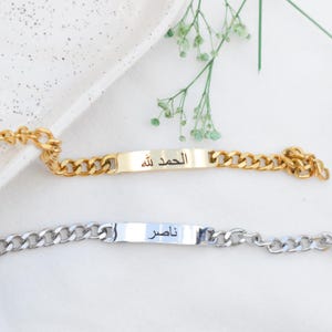 May include: Two metal chain bracelets with rectangular identification plates. The top bracelet is gold-toned and features the Arabic text الحمد لله. The bottom bracelet is silver-toned and features the Arabic text ناصر. Both bracelets have curb links.