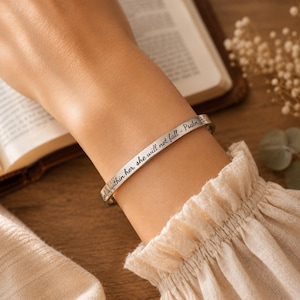 May include: A silver-toned metal cuff bracelet featuring the engraved black text "God is within her, she will not fall - Psalm 46:5". The bracelet has a smooth, polished finish and a slim, open-ended design that wraps around the wrist.