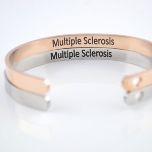 May include: Two open cuff bracelets, one in rose gold tone and one in silver tone, stacked together. Each bracelet features the text Multiple Sclerosis engraved in a clear black font on the outer surface. The ends of the cuffs are rounded.