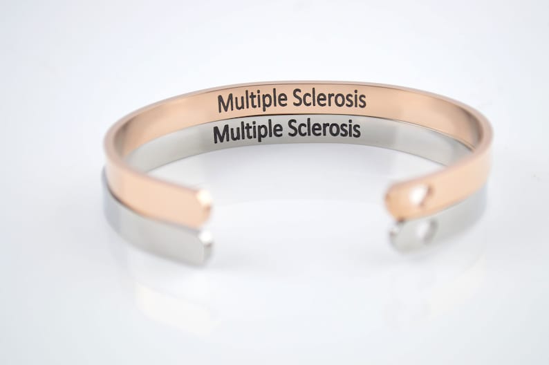 May include: Two open cuff bracelets, one in rose gold tone and one in silver tone, stacked together. Each bracelet features the text Multiple Sclerosis engraved in a clear black font on the outer surface. The ends of the cuffs are rounded.