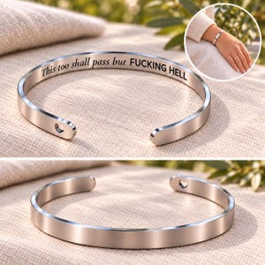 Op de afbeelding: Een zilverkleurige metalen armband met een open ontwerp. De binnenzijde is gegraveerd met de zwarte tekst "This too shall pass but FUCKING HELL". De uiteinden van de armband hebben kleine hartvormige uitsparingen aan de binnenzijde.