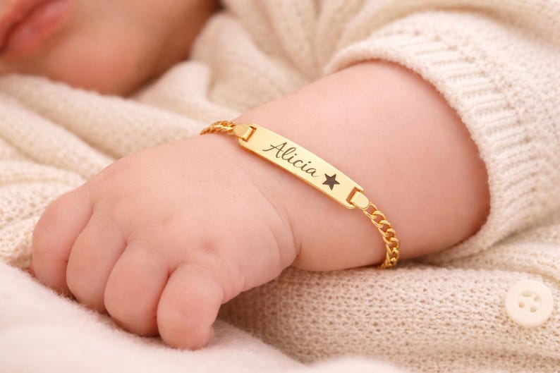 May include: A gold-toned metal identification bracelet featuring a flat rectangular bar engraved with the name Alicia and a small star symbol. The bracelet is constructed with a delicate curb chain link design and a secure clasp mechanism.