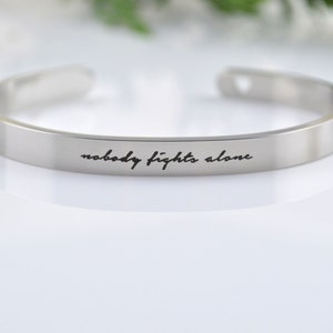 May include: A silver-toned metal cuff bracelet with an open design, featuring the engraved black script text "nobody fights alone" centered on the outer surface of the band. The bracelet has a smooth, polished finish and rounded ends.