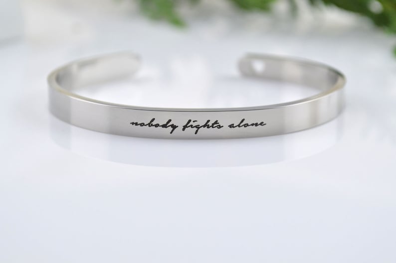 May include: A silver-toned metal cuff bracelet with an open design, featuring the engraved black script text "nobody fights alone" centered on the outer surface of the band. The bracelet has a smooth, polished finish and rounded ends.