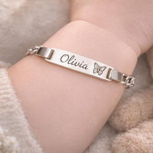 May include: A silver-toned metal identification bracelet featuring a flat rectangular center plate engraved with the name Olivia in a cursive script followed by a small butterfly icon, connected to a polished curb chain link band.