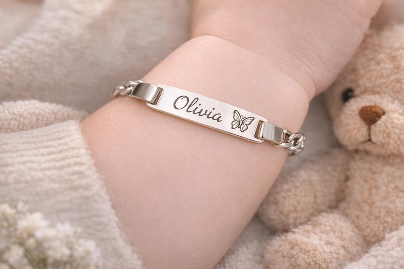 May include: A silver-toned metal identification bracelet featuring a flat rectangular center plate engraved with the name Olivia in a cursive script followed by a small butterfly icon, connected to a polished curb chain link band.