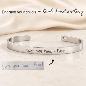 May include: A silver-toned metal cuff bracelet featuring the engraved text 'Love you dad - Ariel' on the outer surface. A small heart cutout is visible at one end of the band. Below the bracelet, a paper note displays the same handwritten text.