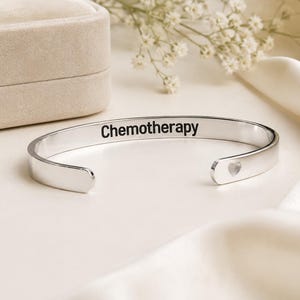 May include: A silver-toned metal cuff bracelet featuring the word Chemotherapy engraved in black lettering on the outer surface. The bracelet has an open design with a small heart-shaped cutout detail visible on one of the rounded ends.