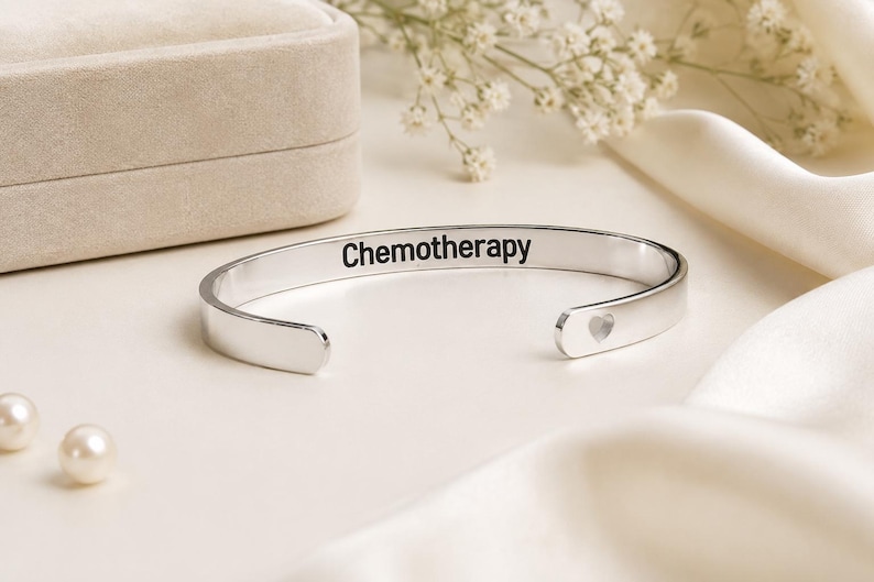 May include: A silver-toned metal cuff bracelet featuring the word Chemotherapy engraved in black lettering on the outer surface. The bracelet has an open design with a small heart-shaped cutout detail visible on one of the rounded ends.