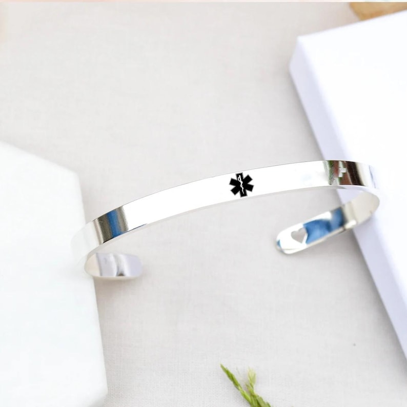 May include: A polished silver-tone metal cuff bracelet featuring a black Star of Life medical alert symbol engraved on the front surface. The open-ended band has a smooth, reflective finish and a small heart-shaped cutout detail at one end.