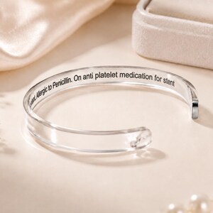 May include: A clear, open-ended cuff bracelet featuring black engraved text on the inner surface that reads: Allergic to Penicillin. On anti platelet medication for stent. The bracelet has a smooth, polished finish and a simple, circular design.