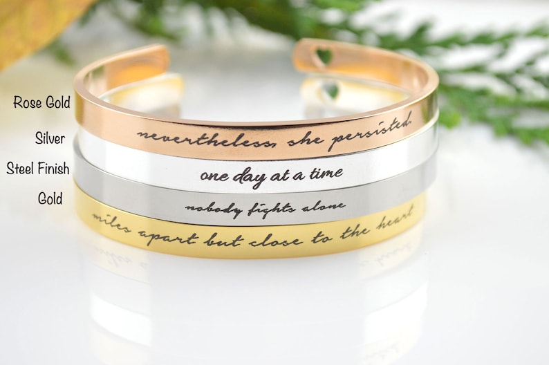 May include: Four metal cuff bracelets stacked vertically, featuring rose gold, silver, steel, and gold finishes. Each bracelet is engraved with black cursive text: nevertheless, she persisted, one day at a time, nobody fights alone, and miles apart but close to the heart.
