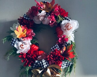 May include: A fall-themed wreath with red, white, and pink flowers, green leaves, and orange pumpkins. The wreath is decorated with black and white checkered ribbon and a gold bow.