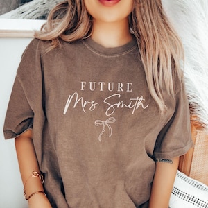 May include: A brown t-shirt with the text "FUTURE Mrs. Smith" in white lettering with a small bow underneath.