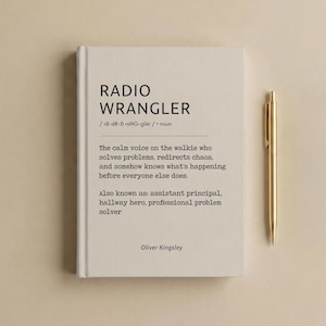 May include: A cream-colored hardcover notebook with black text reading RADIO WRANGLER, a phonetic pronunciation, and a definition describing a calm voice on a walkie-talkie. A gold-toned metal pen is positioned vertically to the right of the notebook.