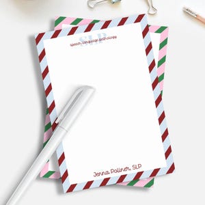 May include: Two white notepads with a diagonal striped border in red, blue, and green. The text "SLP speech language pathology" and "Jenna Poliner, SLP" are printed on the notepad. A white pen rests on the notepad.