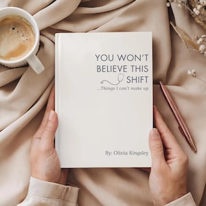 May include: A white book titled YOU WON'T BELIEVE THIS SHIFT ...Things I can't make up by Olivia Kingsley, featuring a small stethoscope graphic. The book is held in place with a cup of coffee and a rose gold pen nearby on a soft fabric surface.