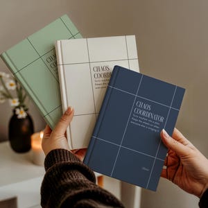 May include: Three hardcover journals in sage green, off-white, and navy blue, each featuring a grid pattern and the text "CHAOS COORDINATOR. Turns mayhem into a plan. Keeps the wheels on when everyone else is winging it." The navy journal includes the name "Oliver."