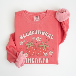May include: A pink sweatshirt with the text "Occupational Therapy" in a retro font, with two strawberries and flowers printed on the front.