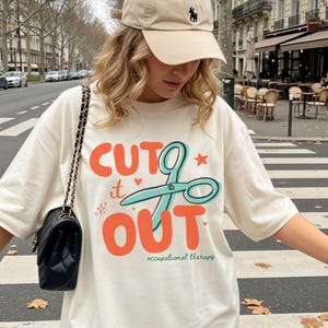 May include: A cream-colored t-shirt featuring the orange text CUT IT OUT with a teal graphic of scissors, small orange hearts, and stars. Below the text, the words occupational therapy are printed in small green letters. A black quilted shoulder bag.
