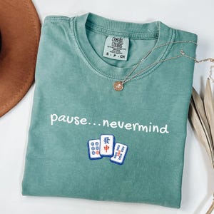 May include: A sage green short-sleeve t-shirt with a crew neckline. The shirt features the white text "pause...nevermind" above a graphic of three mahjong tiles. A gold-toned necklace with a small circular pendant rests on the collar area.
