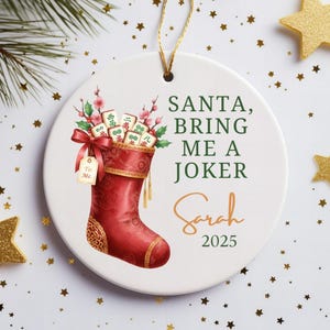 May include: A round white ceramic ornament featuring a red Christmas stocking filled with mahjong tiles and pink flowers. The text reads SANTA, BRING ME A JOKER, Sarah, and 2025. A gold string is attached to the top for hanging purposes.