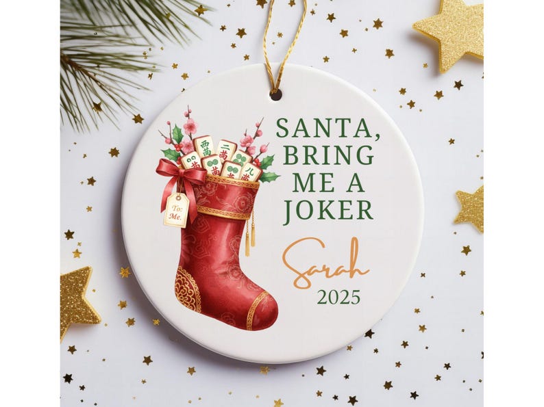 May include: A round white ceramic ornament featuring a red Christmas stocking filled with mahjong tiles and pink flowers. The text reads SANTA, BRING ME A JOKER, Sarah, and 2025. A gold string is attached to the top for hanging purposes.
